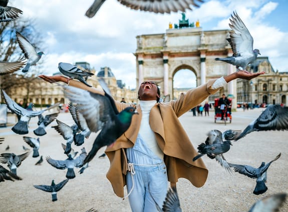 young female standing in the middle of pigeons taking off in a city square
