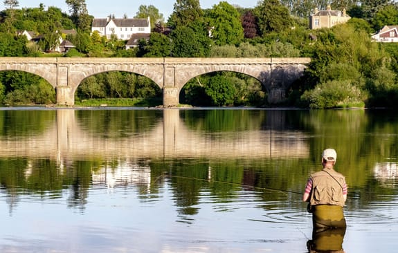 Fisherman wades waste-deep in river in foreground of an arched bridge