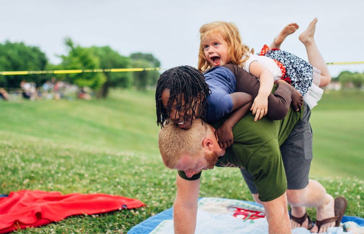 Man on picnic rug bears the weight of two young children jumping playfully on his back