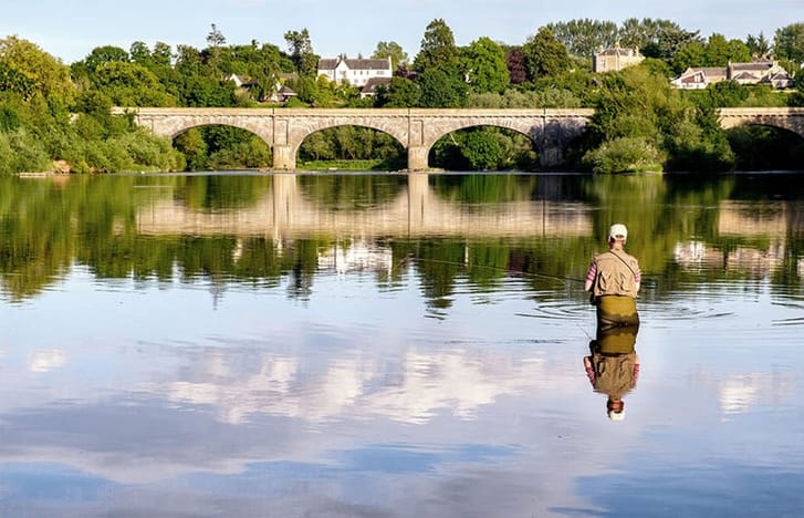 Fisherman wades waste-deep in river in foreground of an arched bridge
