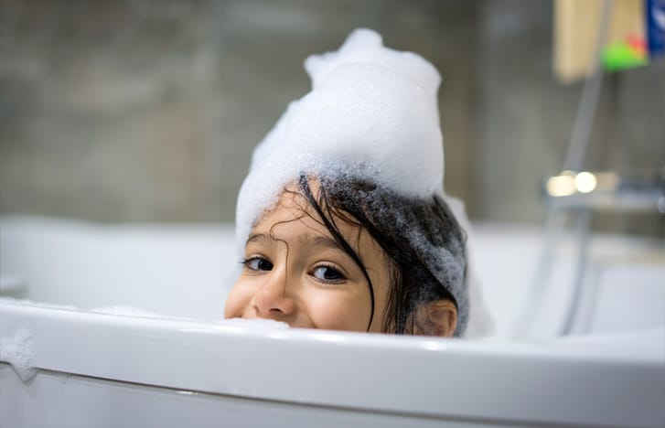 Young girl covered in suds cheekily peeks over bathtub