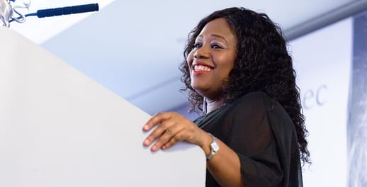 A lady smiles as she speaks at a lectern to a crowd