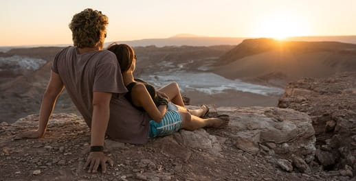 Young couple sitting on top of mountain rocks looking out towards skyline and sunset