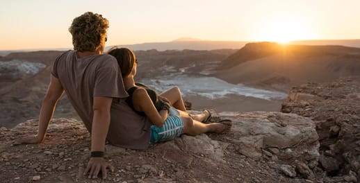 Young couple sitting on top of mountain rocks looking out towards skyline and sunset