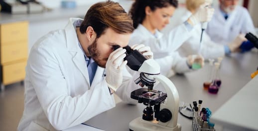A scientific researcher in a laboratory coat looks into a microscope surrounded by colleagues