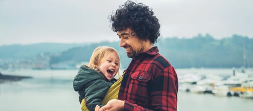 A man holds his child in a baby carrier while at the seaside