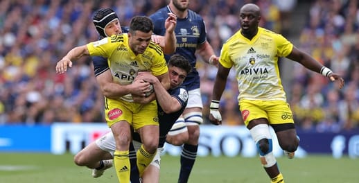A rugby player staggers towards the try line while being tackled by opponents
