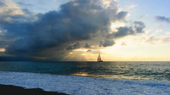 sailing ship in the distance with sun breaking through stormy clouds