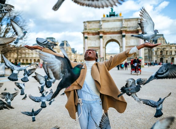 Female standing in a square with pigeons flying around her