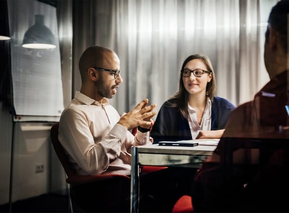 A man is folding his hands while talking during a meeting, as a woman next to him is listening and smiling