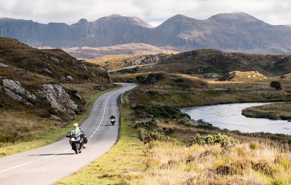 Two motorbikes riding along a road next to a rive in the Scottish Highlands