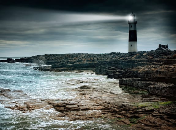 lighthouse surrounded by rocks with light on guiding the ships