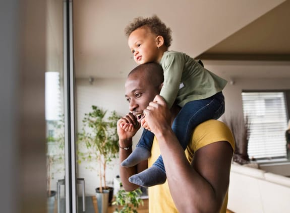 A man holds a toddler on his shoulders at home