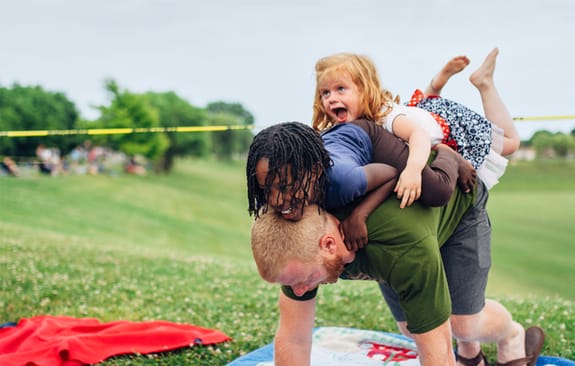 Man on picnic rug bears the weight of two young children jumping playfully on his back