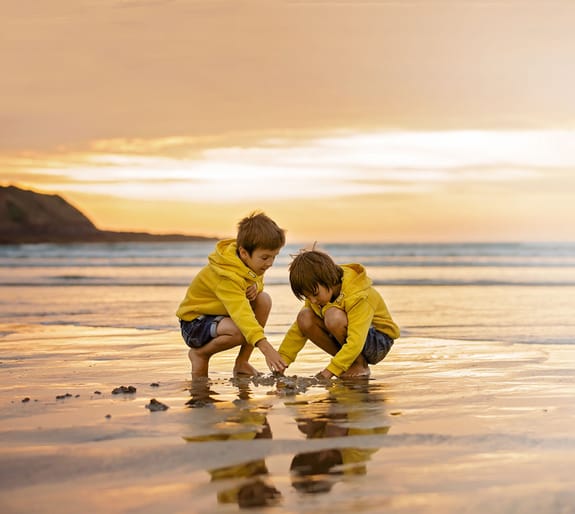 Two young boys, brothers, play at the beach at sunset. A focus is on the horizon.
