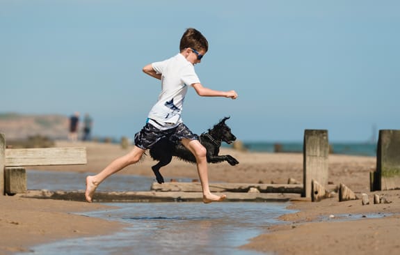 young boy and his dog jumping over a puddle on the beach