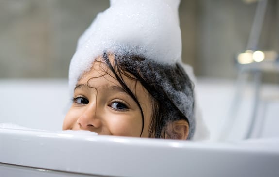 Young girl covered in suds cheekily peeks over bathtub