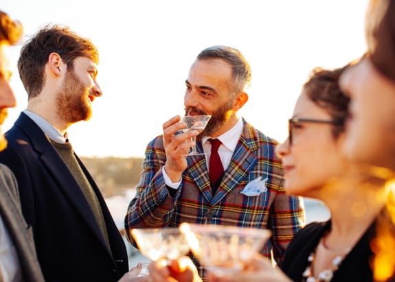 A middle-aged man in tartan suit sips a martini at a country event while talking to a guest