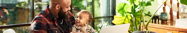 Father sitting with child on his lap while working on laptop at kitchen table
