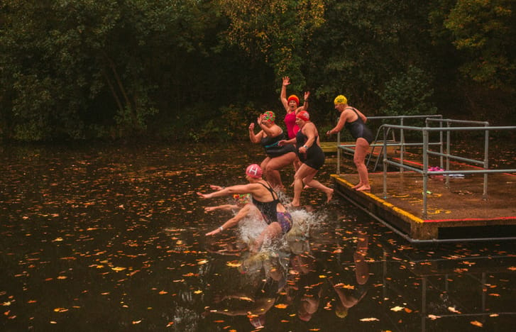 Six older women in swimming kits bravely jumping into a cold autumn pond