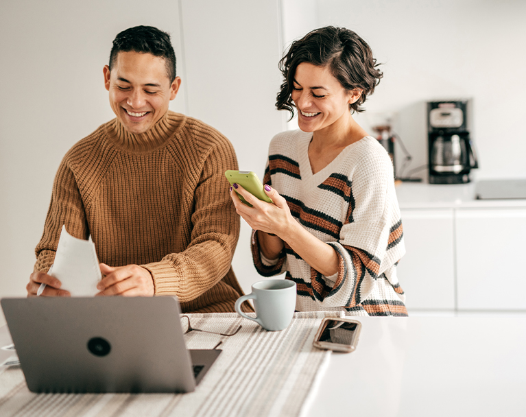 Smiling couple at kitchen table checking laptop and phone