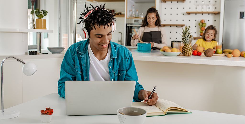 Investec Youth Account: Male teenager smiling taking notes in front of his laptop while his sisters are in the kitchen