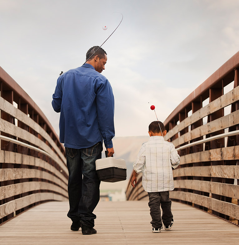 Father walking along a wooden bridge with his son, both carrying fishing rods