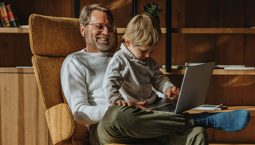 Older male sitting in a rocking chair with his grandson looking at a laptop