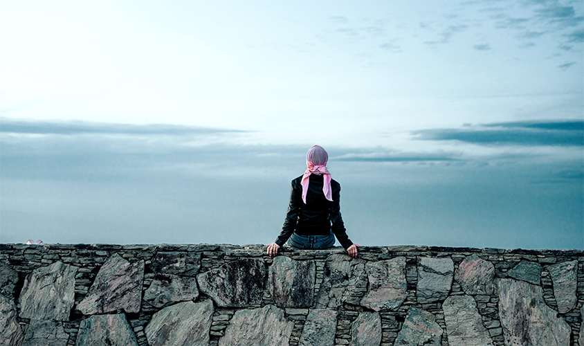 Female wearing a head scalf sitting on a stone wall looking out over the ocean