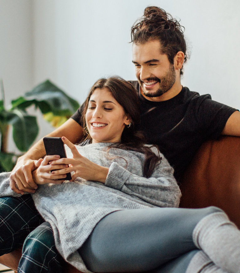 A couple relaxing on a couch in their first-time home in South Africa, with the woman holding a smartphone.