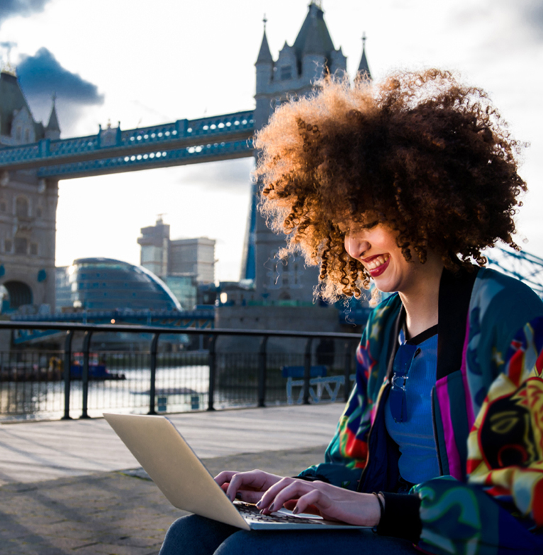 A young woman manages her offshore account on a laptop outdoors, with London's Tower Bridge in backdrop
