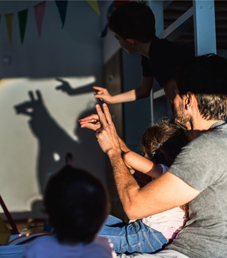 A family enjoying a shadow puppet play created by hand silhouettes against a bright wall while discussing their family bank account.