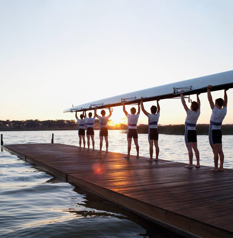 A team of rowers carry their rowing boat upside down towards the water at sunrise, in readiness to go rowing