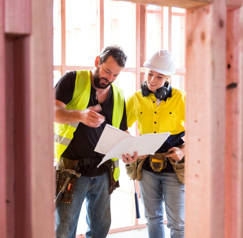 Two builders look a set of house drawings while standing amongst a new house's timber frame