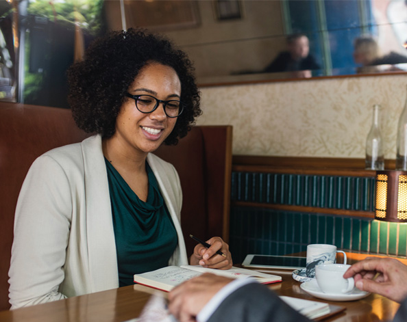 A lady wearing a beige blazer takes notes in a notebook while having coffee with someone