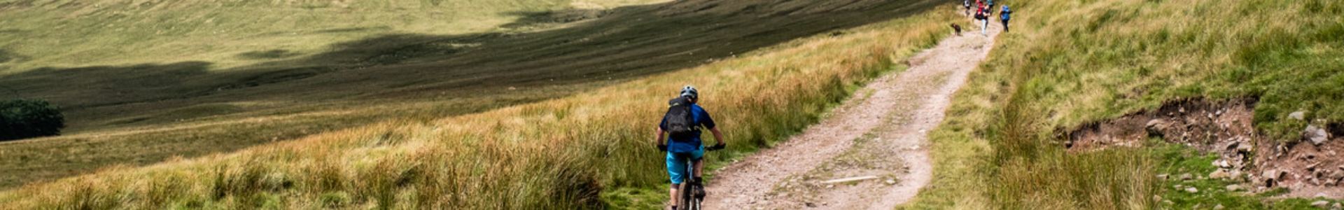 A loan mountain biker rides up a path in a grassy hill in the Brecon Beacons, as a group of runners descend towards him