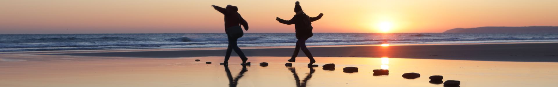 A couple hop across stones on the sand at a beach while the sun sets on an English winter's day