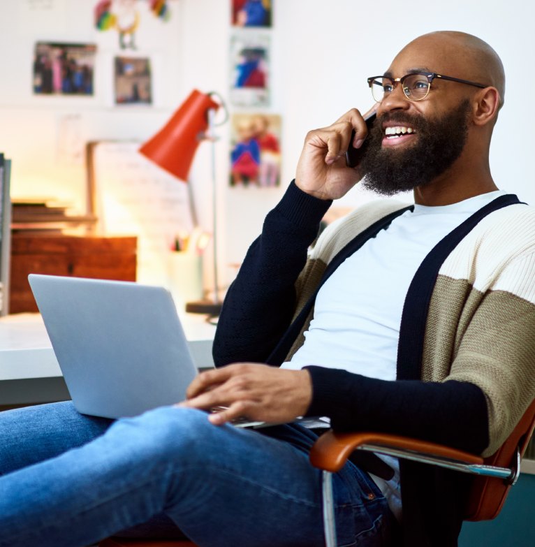 A man leans back in a chair with a laptop on his lap while speaking on the phone